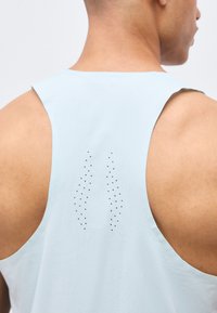 Κοντινή λήψη του πάνω μέρους της πλάτης ενός ατόμου που φοράει αθλητική μπλούζα χωρίς μανίκια σε ανοιχτό μπλε χρώμα με οπές αερισμού σε δύο κάθετα μοτίβα.