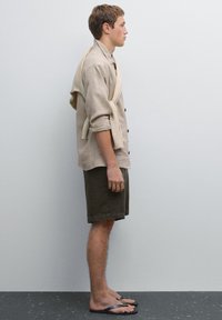 Light beige linen shirt with rolled sleeves, paired with dark green shorts, and black flip-flops. A tan sweater drapes over the shoulders.
