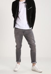 Giacca in suede nera con spalle rigate, t-shirt grafica bianca, jeans grigi e sneakers bianche. Outfit casual con texture sovrapposte.