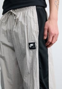 Světlé šedé kalhoty s elastickým pasem a stahovací šňůrkou. Mají texturovanou látku s černým síťovaným bočním panelem a nášivkou s logem Nike Air.