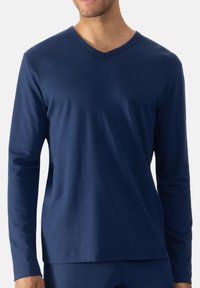 Man in een strakke, langmouwige, donkerblauwe V-hals shirt met een gladde textuur tegen een effen lichte achtergrond.