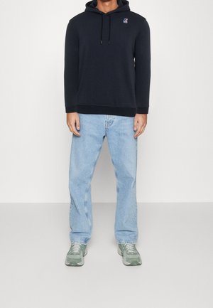 Uomo con felpa nera con logo, jeans azzurri chiari e scarpe da ginnastica verdi e bianche, in piedi su un pavimento uniforme contro uno sfondo bianco.