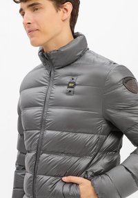 Grau gepolstertes Jacket mit hohem Kragen, gesteppter Gestaltung und zwei sichtbaren Fronttaschen. Verfügt über einen Logo-Patch auf der Schulter und einen Reißverschluss.