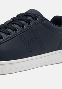 Zapatillas de tela sintética de color azul marino con cordones redondos, suela de goma blanca y un sutil logo en forma de 'H' perforado en el lateral.