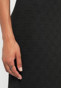 Svart texturerad klänning med ett präglat mönster av varumärket, med en närbild av tyget och en hand placerad nära fållen.
