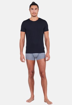 Mand stående barfodet iført en sort t-shirt med korte ærmer og grå boxershorts mod en ensfarvet lys baggrund.