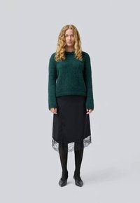 Green textured sweater and black knee-length skirt with lace hem, paired with black tights and shoes, model standing against a neutral background.