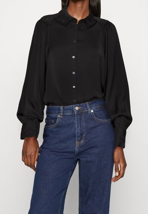 Femme portant un chemisier noir à manches longues boutonné avec des manches bouffantes, associé à un jean bleu foncé taille haute.