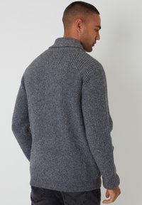 Szary sweter z dzianiny o prążkowanej strukturze, złożonym kołnierzem i długimi rękawami, widziany od tyłu na jasnym tle.