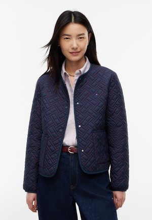 Femme aux cheveux raides et foncés porte une veste matelassée bleu marine, une chemise rayée boutonnée, un jean foncé et une ceinture marron sur fond blanc.