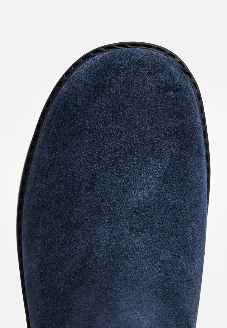 Bottine en suede bleu marine avec une texture lisse, bout rond et semelle en caoutchouc noir contrastante. Pas de quincaillerie ni d'accents visibles.