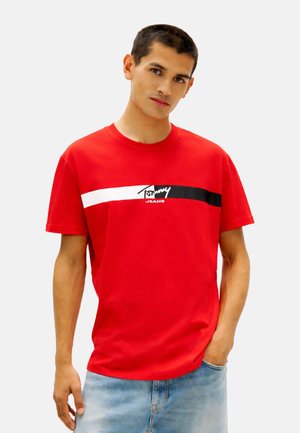 Joven con el pelo corto y oscuro, vestido con una camiseta roja de Tommy Jeans y vaqueros azul claro, de pie con una mano en el bolsillo.