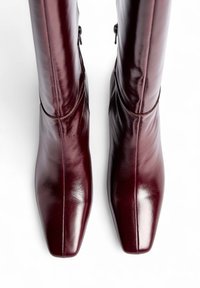Burgundijas ādas ceļgala augstuma zābaki ar gludu tekstūru, kvadrātveida purngaliem un plānu izšuvumu, ar sānu rāvējslēdzēju slēdzēšanai.