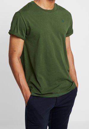 Mann trägt ein olivgrünes, kurzärmeliges T-Shirt mit hochgekrempelten Ärmeln und dunkelblaue Hose, Hände in den Taschen, vor einfachem hellem Hintergrund.