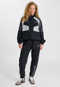 Must hall komplekt hallide detailidega; lukuga jakk, millel on kõrge krae ja elastsed mansetid, koos kitsenevate pükstega. Rinnal on tähelepanuväärne brändi logo.