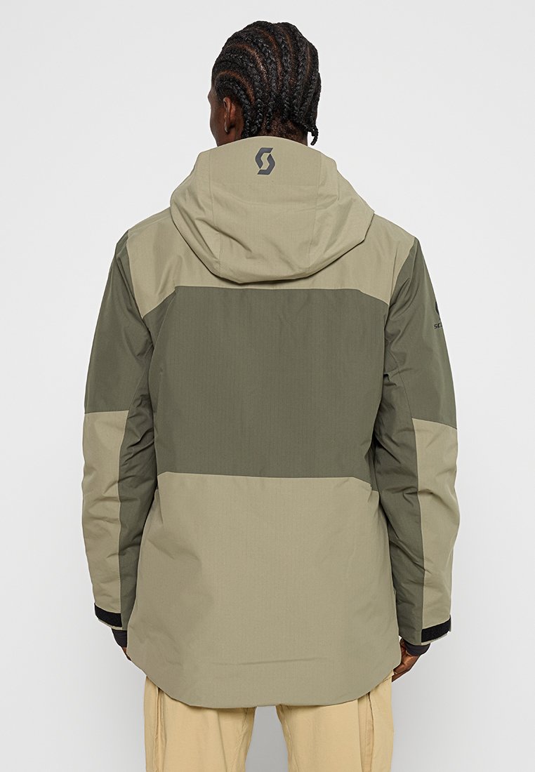 Persona con cabello trenzado que lleva una chaqueta con capucha verde bicolor y pantalones beige, vista desde atrás contra un fondo liso.