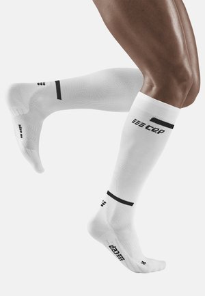 Weiße knielange Kompressionssocken mit schwarzen Akzenten und Logo, ausgestattet mit einer strukturierten Oberfläche und Stützbanden nahe dem oberen Rand und dem Fußgewölbe.