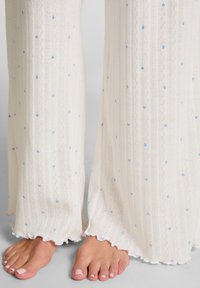 Witte gebreide broek met een tekstuurpatroon, voorzien van kleine blauwe hartjes. De randen hebben een golvende snit. Zichtbare blote voeten met lichte nagellak.