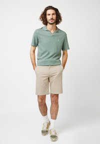 Polo shirt verde de manga corta con cuello plano, pantalones cortos beige y zapatillas con detalles en verde y naranja. Diseño simple y casual.
