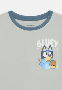 T-shirt de algodão cinza claro com gola redonda azul. Apresenta uma personagem impressa a segurar um gelado, rotulada com a palavra "BLUEY."