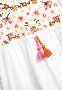 Valkoinen vaate, jossa on värikästä kukka-ompelua vaaleanpunaisena, oranssina ja keltaisena. Sisältää vaaleanpunaiset ja oranssit hapsut helman kohdalla.