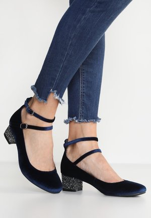 Voeten met donkerblauwe fluwelen enkellaarsjes met glinsterende blokhakken en versleten donkerblauwe skinny jeans op een witte achtergrond.