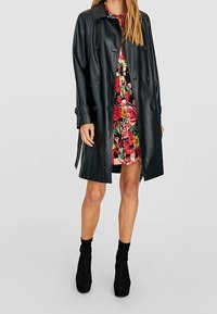 Svart läder trench coat med dubbelknappring, bred krage och sidofickor, bärs över en blommig klänning och ankelskor.