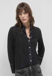 Camicia nera con chiusura a bottoni, dettagli in cucitura bianca, maniche lunghe e colletto. Indossata con pantaloni in denim grigi.