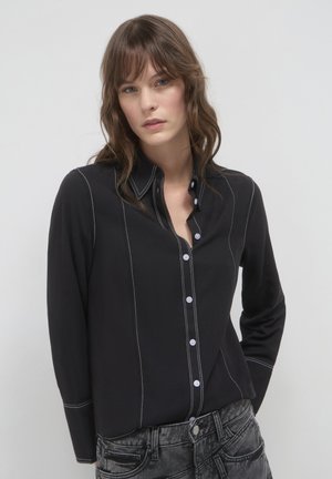 Schwarzes Hemd mit Knopfleiste, weißen Nähten, langen Ärmeln und Kragen. Trägt dazu graue Jeans.