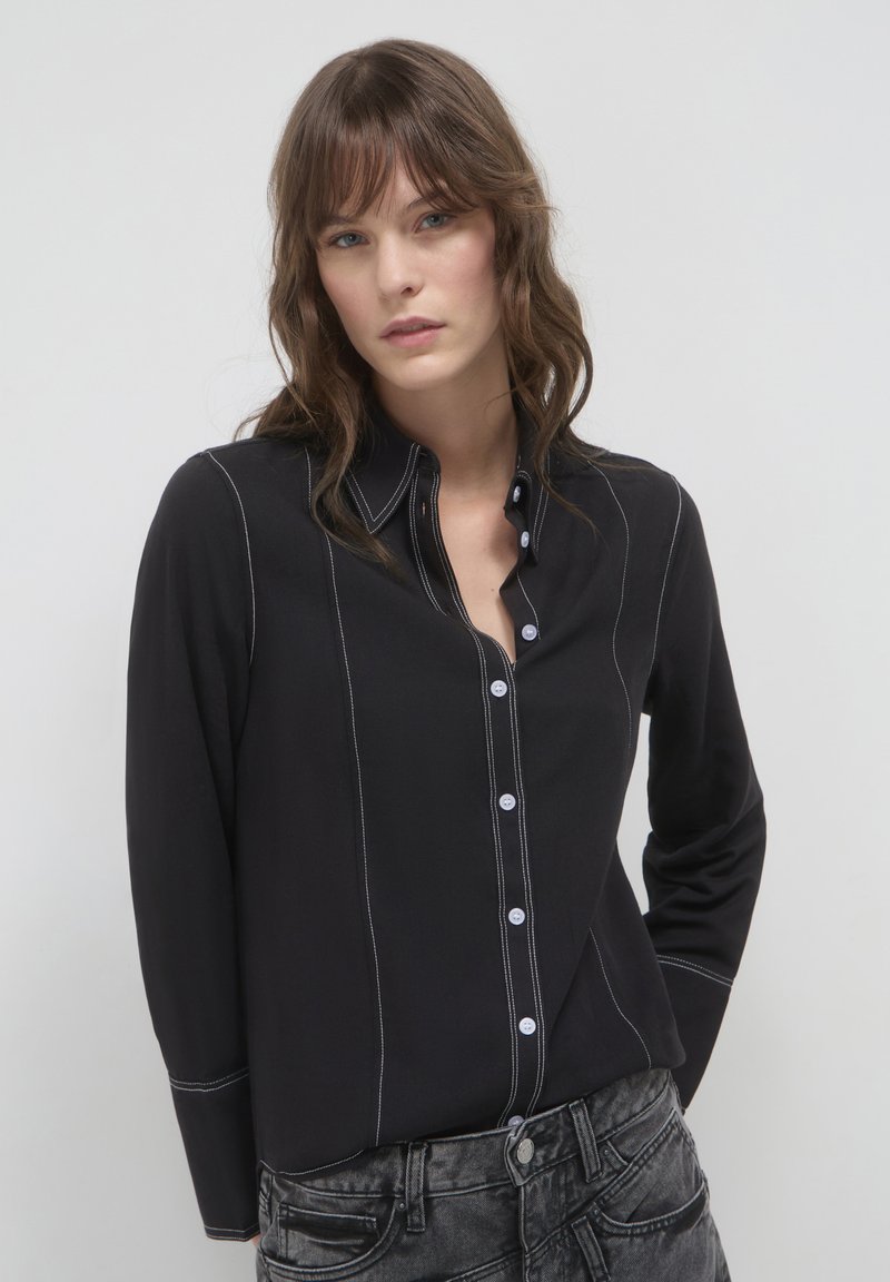 Camicia nera con chiusura a bottoni, dettagli in cucitura bianca, maniche lunghe e colletto. Indossata con pantaloni in denim grigi.