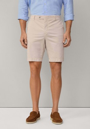 Homme portant un short beige longueur genoux, une chemise bleu clair à manches longues retroussées et des chaussures marron sans lacets, debout sur un fond uni.