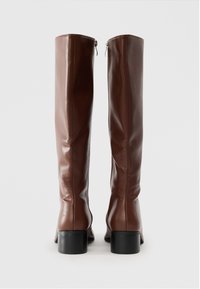 Une paire de bottes en cuir marron haute avec des talons blocs et des zips à l'arrière sur un fond blanc uni.