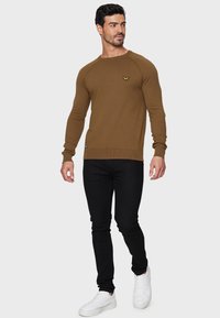 Brun langærmet striktrøje med ribbede manchetter og kant, prydet med et lille logo på brystet, kombineret med sorte slim-fit jeans og hvide sneakers.