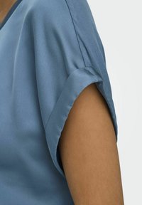 Top corto blu satinato a maniche corte con risvolto, morbido al tatto e design a V, che mostra parte del braccio su uno sfondo neutro.