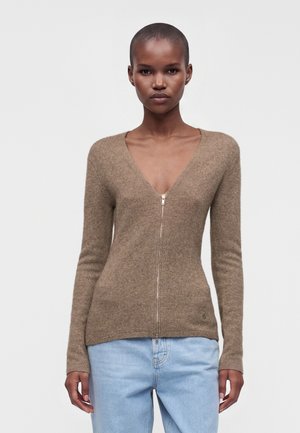 Femme portant un cardigan marron ajusté zippé et un jean bleu clair, se tenant devant un fond gris clair uni.