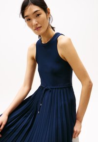 Rochie plisată bleumarin cu un corset ajustat din material canalat, decolteu rotund și talie cu șnur reglabil. Țesătură moale cu o textură subtilă.