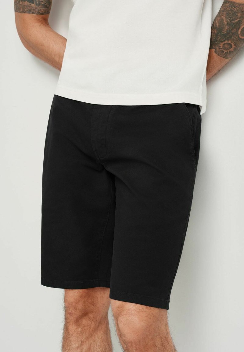Czarne bawełniane szorty o luźnym kroju, z przednimi kieszeniami i gładką fakturą. Stylizowane z prostą białą koszulą u góry.