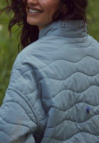 Glunderende vrouw met krullend haar draagt een lichtblauwe gewatteerde jas, met een klein blauw bloemetje in de stof gestoken, buiten met een vage groene achtergrond.
