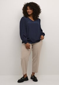 Blusa de color azul marino, de manga larga, con escote en V y patrón texturizado, combinada con pantalones beige y mocasines negros. La blusa tiene puños fruncidos.