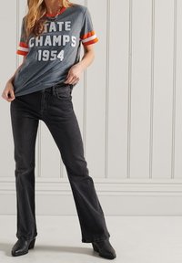 Camiseta de algodón gris con "STATE CHAMPS 1954" en letra blanca y mangas a rayas naranjas. Combinada con jeans de mezclilla negros de pierna acampanada y botas de tobillo.