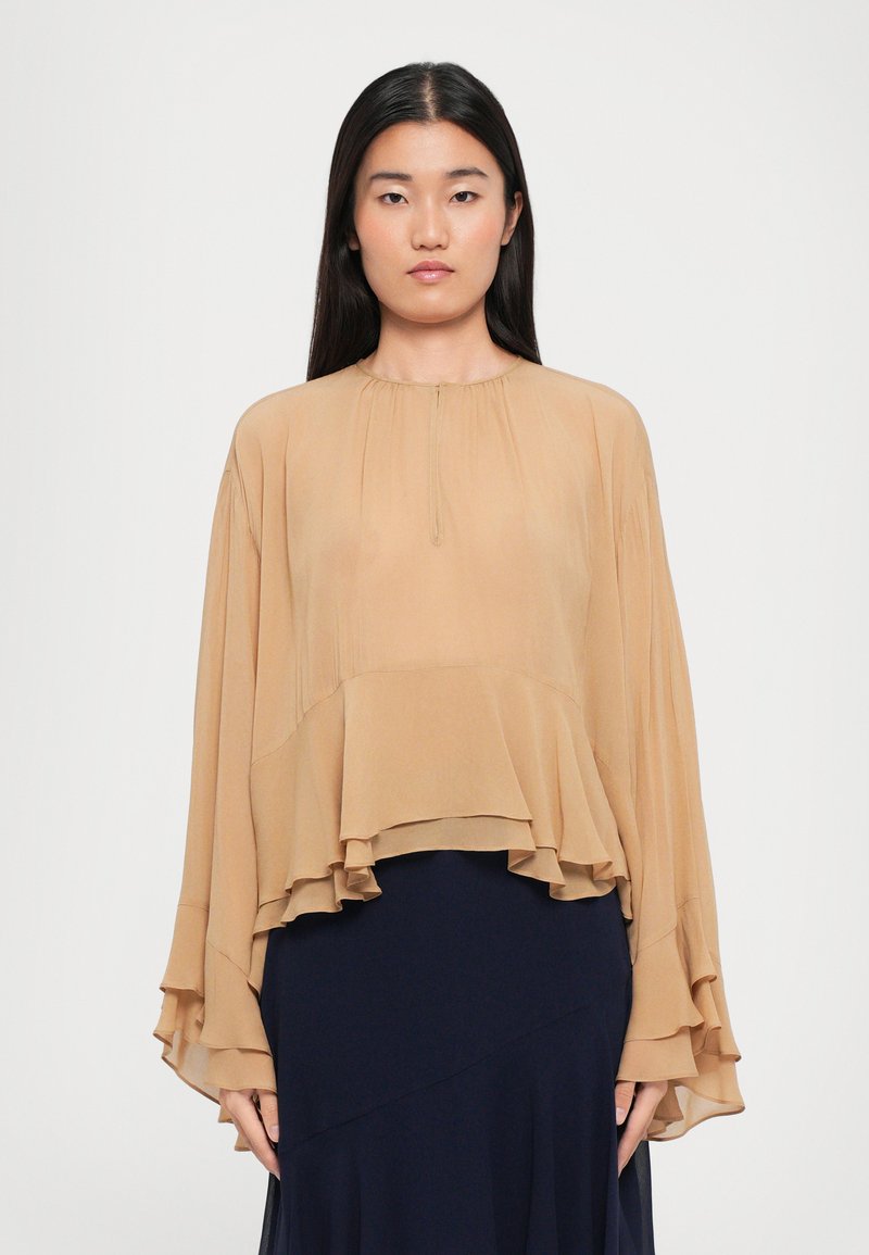 Kvinde iført en løs beige lagdelt bluse med brede ærmer og en mørk marineblå nederdel, stående foran en ensfarvet lys baggrund.