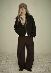 Crna bomber jakna s kapuljačom obloženom krznom, smeđi pleteni pulover ispod i široke crne hlače s asimetričnim dizajnom. Crna obuća.