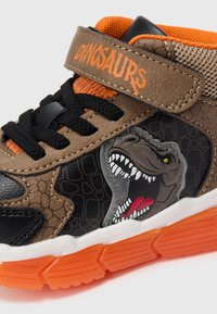 Adidași sport în nuanțe de marron și negru cu grafică de dinozaur, accente portocalii, panouri texturate și șireturi negre. Curea Velcro etichetată "DINOZAURI".