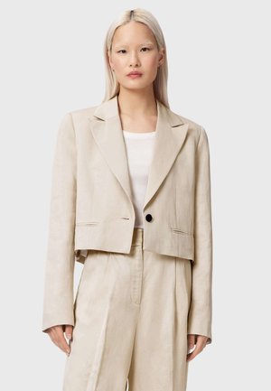 Kvinde med lige blondt hår iført en beige cropped blazer og matchende højtaljede bukser over en hvid top, stående mod en ensfarvet baggrund.