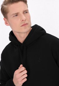 Sort hætte sweatshirt med en struktureret stof, en snørehætte og et diskret ankerlogo på brystet. Nærbillede af en mandlig model.