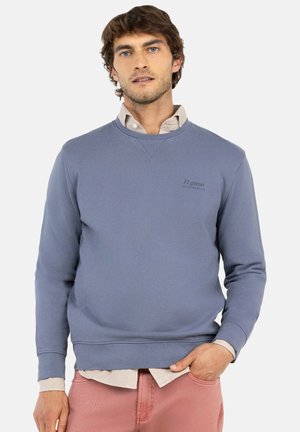 Hombre con sudadera azul claro sobre camisa beige y pantalones rosas, de pie con una mano en el bolsillo frente a un fondo blanco.