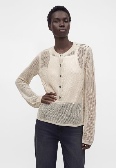 Femme portant un top en maille beige à manches longues et boutonné avec un pantalon foncé, posant avec une main sur la hanche sur un fond clair uni.
