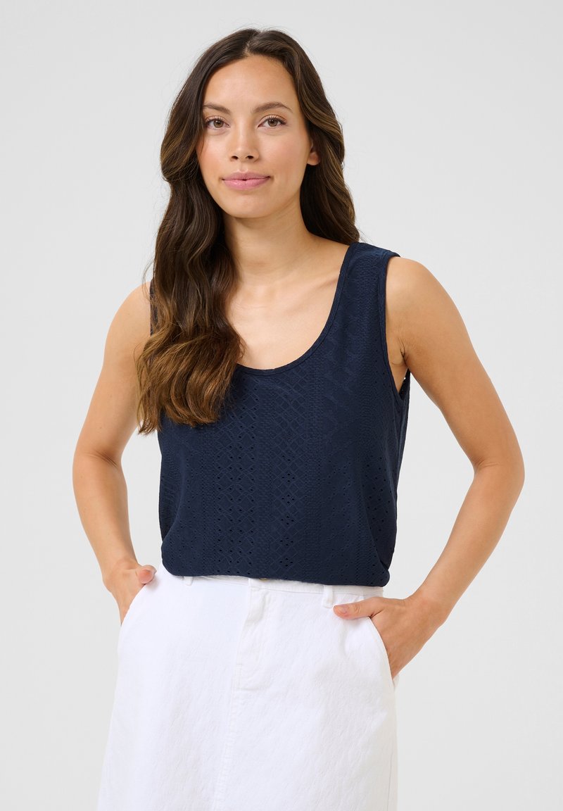 Haut sans manches bleu marine avec un motif texturé de diamants et de lignes, doté d'un décolleté arrondi et d'une coupe décontractée. Associé à une jupe blanche.