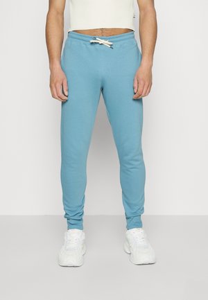 Personne portant un pantalon de survêtement à cordon de serrage bleu clair et des baskets blanches épaisses, debout devant un fond gris clair uni.