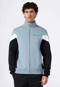 Sudadera con cremallera de color azul claro con acentos en negro y blanco. Fabricada en una tela suave, cuenta con un cuello alto y un logo bordado en el pecho.